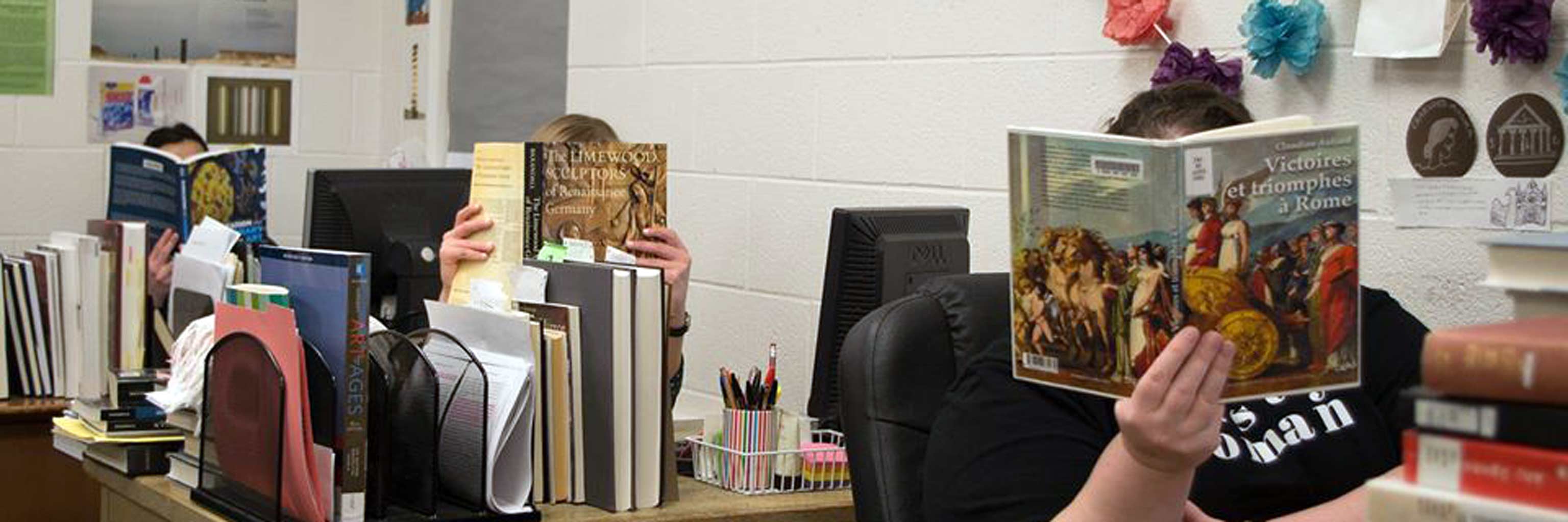 Three students sitting at their desks hiding their faces behind Art History books.